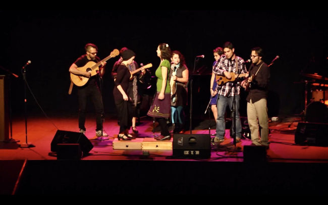 Image of a darkened stage with a folk band of six with various stringed instruments and 2 clog dancers.